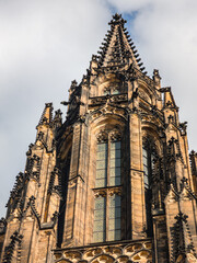 Fototapeta premium Detailed vertical view of a gothic cathedral spire with intricate stone work and gothic windows reaching towards a clear sky with soft white clouds.