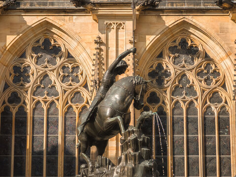 Close up of a historical bronze statue of a knight on a horse in front of a gothic cathedral wall with intricate stone window traceries in daylight.