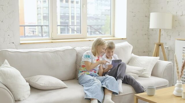 9 years old girl watches as the boy plays a game on his tablet while sitting on sofa at home