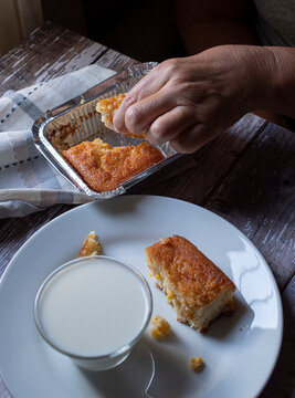 Manipulacion de alimentos, manejo de comida en la cocina, manos tomando un pedazo de pastel de elote de  una charola de aluminio con un vaso de leche por un lado, sobre una mesa de madera y un plato b