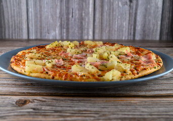Warm pizza featuring ham and pineapple sits on a blue plate. The wooden table background adds a rustic touch. Ideal for casual dining or sharing with friends