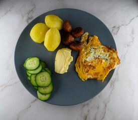 A well-prepared breakfast features a golden fried egg, boiled potatoes, meat balls, fresh cucumber slices, and a dollop of butter on a stylish gray plate