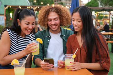 Three friends are enjoying drinks and looking at a smartphone at an outdoor bar