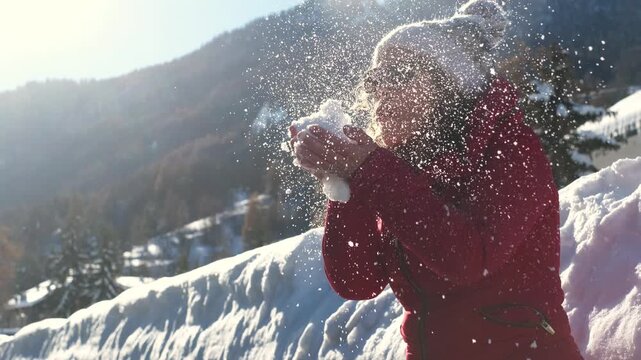 Woman blowing on a small snowball in her hands during a sunny winter day in the mountains, dressed in winter jacket and wool hat, symbol of fun, leisure, vacation, snow and outdoor joy