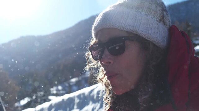 Woman blowing on a small snowball in her hands during a sunny winter day in the mountains, dressed in winter jacket and wool hat, symbol of fun, leisure, vacation, snow and outdoor joy