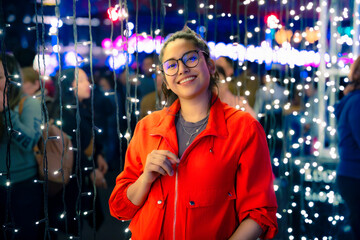 A young smiling Latin woman stands surrounded by twinkling Christmas lights during a festive night...