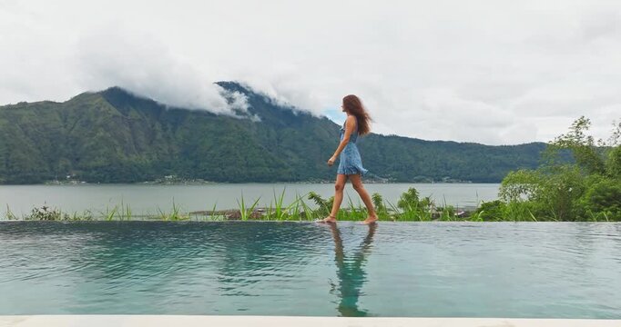 Relax and travel to Bali. Beautiful young woman in a flowing blue dress walks barefoot along the infinity pool edge, overlooking Lake Batur and verdant Batur volcano, panoramic vacation scene