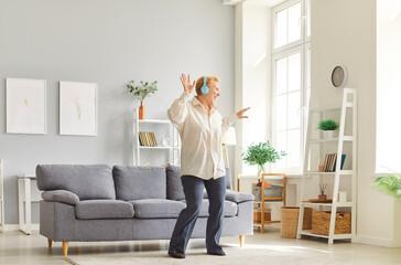 Cheerful mature woman with blonde hair wearing headphones dancing joyfully in bright modern living...