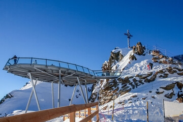 A winter mountain observation deck with a cross on a snow-capped peak under a blue sky
