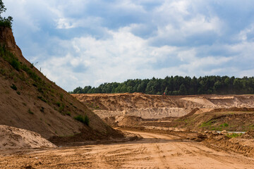 Expansive sand quarry with prominent sandy slope and rough dirt road. Excavator works on distant sandhills, a forest line and cloudy sky complete the scene
