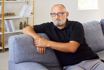 Senior man on sofa at home, thoughtful portrait. Wearing glasses, the mature person rests, a casual...