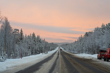 Wide view of a divided highway covered with snow and ice, bordered by a snow-laden forest, extending to the horizon under a pink and orange sky