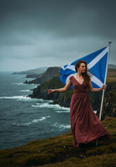 Woman with the Scottish flag stands on a dramatic cliff overlooking the sea. Vertical photo of the rugged Scotland coast. National pride and heritage concept