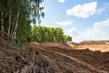 Fototapeta premium Rough brown soil with deep tire tracks dominates the foreground, hinting at ongoing excavation near a vibrant green forest and distant sand quarry under a clear blue sky
