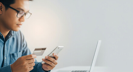Young man holding credit card and smartphone while shopping online, sitting at desk with laptop, focused and calm, modern technology and digital payment concept