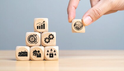 Wooden blocks with business icons stacked in pyramid shape. A hand places another block
