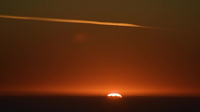 A golden sunset over the dramatic landscape of Wadi Shab and the tranquil Village Al Fahal in Oman. The sky glows with vibrant orange and red hues, with a single contrail streaking across the vast exp