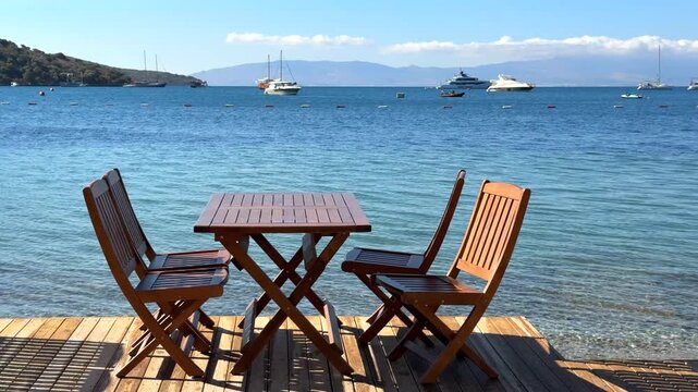 Seaside dining deck with wooden tables and chairs overlooking a beautiful turquoise bay, sailboats, and distant hills under a clear blue sky.