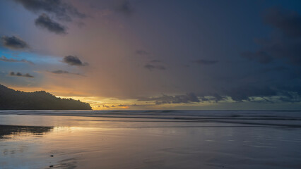 Beautiful tropical sunset. Ocean waves are spreading across the sandy beach. Reflection on wet, smooth sand. Purple clouds in the sky, illuminated by orange near the horizon. Long exposure. Malaysia. 