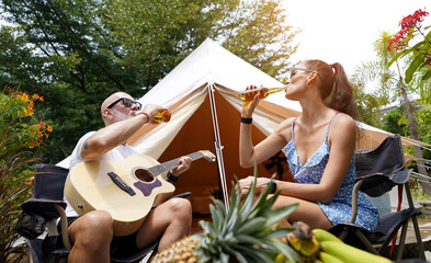 A man and woman is joyfully drinking beer and playing guitar near a cozy tent at glamping