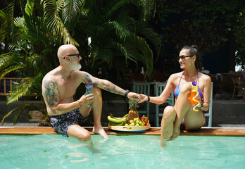 A man and a woman sit by the pool with cocktails and fruit.
