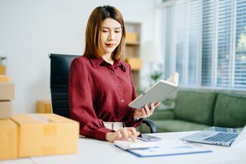 Startup small business SME, Entrepreneur owner woman using smartphone or tablet taking receive and checking online purchase shopping order to preparing pack product box.