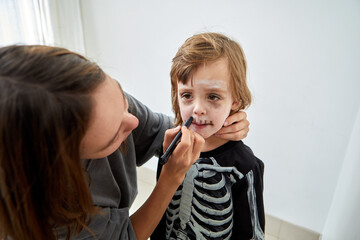 A young child, cheerfully dressed in a skeleton outfit, happily receives colorful makeup for a spooky Halloween look
