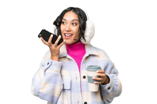 Young Argentinian woman wearing winter muffs over isolated background holding coffee to take away and a mobile