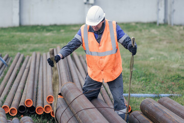A construction worker in an orange vest and helmet is managing pipes at a construction site,...