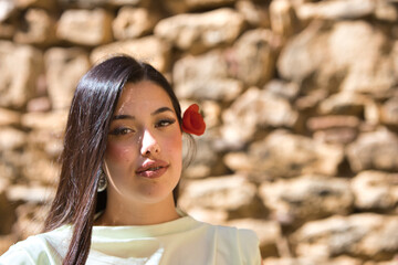 A beautiful Spanish teenager with long dark hair and a delicate, beautiful red poppy in her hair next to her face. The woman is happy and passionate. In the background is a stone wall.
