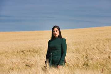 A beautiful, dark-haired teenage Latin girl dressed in green stands among a dry, yellow wheat field on a summer day. In the background, the blue sky.