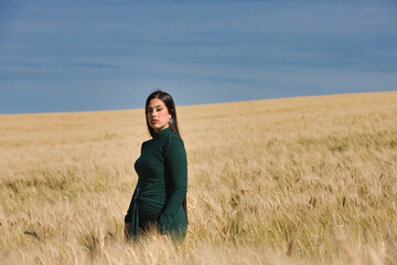 A beautiful, dark-haired teenage Latin girl dressed in green stands among a dry, yellow wheat field on a summer day. In the background, the blue sky.