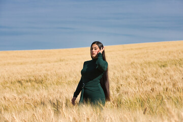 A beautiful, dark-haired teenage Latin girl dressed in green stands among a dry, yellow wheat field on a summer day. In the background, the blue sky.