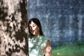 A beautiful Latin teenager with long dark hair rests her hands and face on a tree trunk in the sunlight. The woman is sad and depressed and looks in different directions.