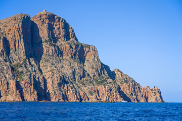 Naklejka premium cap rosso with dramatic red steep cliffs and genoese tower in the Gulf of Porto seen from the sea under blue skies and sunlight