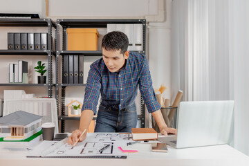 A young architect works with building plans and a house model in the office, reviewing blueprints, marking details, and analyzing design structures with focus and creativity.