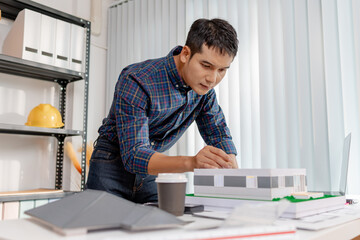 A young architect works with building plans and a house model in the office, reviewing blueprints, marking details, and analyzing design structures with focus and creativity.