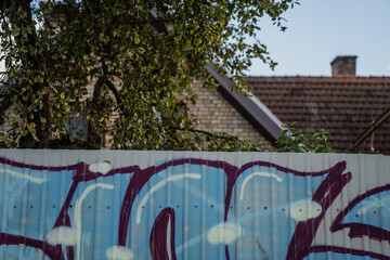Corrugated metal fence with colorful graffiti art in front of brick house and tiled roof showing contrast between urban expression, suburban architecture and contemporary visual culture