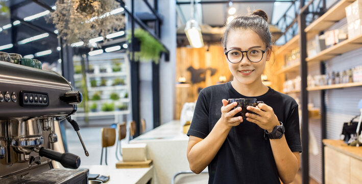 Smiling young Asian woman barista wearing glasses and black shirt holds a coffee cup, standing in front of a coffee machine in her modern cafe - Powered by Adobe