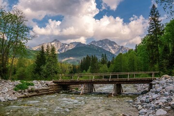 Fototapeta premium Peaceful mountain landscape with a wooden bridge over a clear stream, surrounded by lush green forest and majestic peaks under a partly cloudy blue sky in spring or early summer.