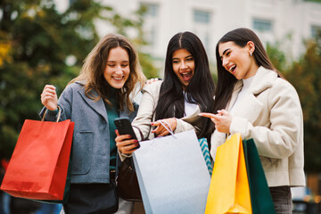Three young women smiling, holding shopping bags, and looking at a smartphone outdoors
