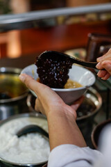 Hand serving traditional Indonesian black glutinous rice porridge, known as bubur ketan hitam, into a bowl with coconut milk. Authentic Asian dessert scene showing local food culture