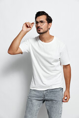Fototapeta premium Thoughtful man adjusting glasses while looking into the distance. He wears a casual white t-shirt and blue jeans against a plain light background. Personal expression concept.