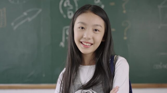 Back to school concept, portrait of a happy Asian child girl student in a uniform with a school bag backpack with a blackboard standing smiling and arms crossed while looking at camera in a classroom.