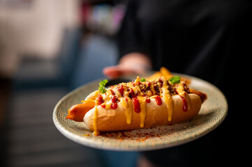 Close-up of a hot dog with mustard, ketchup, onions, and parsley on a patterned plate. Garnished with seasoning. Blurred background. Fast food concept, vibrant and appetizing.