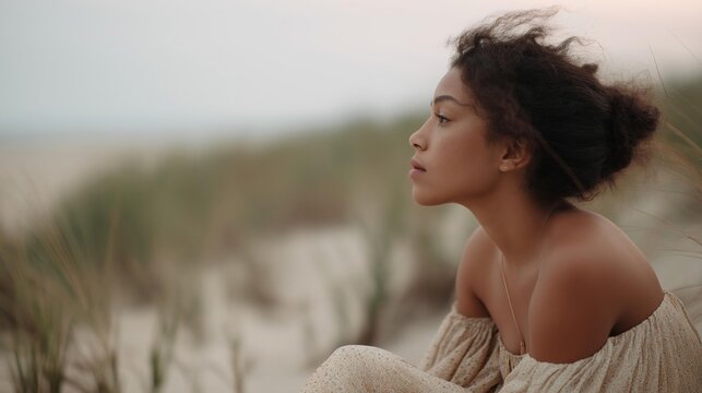 Portrait of a young woman sitting on a sandy beach. she is wearing a beige off-the-shoulder dress with a floral pattern and her hair is styled in loose curls.