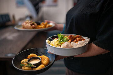 Waiter holding three plates with fried shrimp, curly fries, chicken wings, and a hot dog in a casual restaurant setting. Fast food presentation, handheld service, vibrant dining atmosphere.
