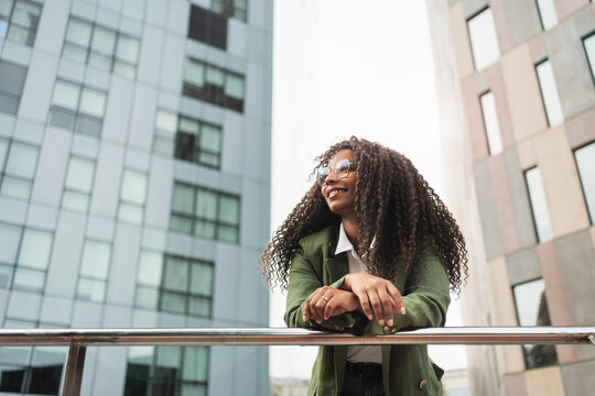 Young African American business woman in trendy outfit and glasses standing in corporate urban district. Inspired creative female professional looking away in the city