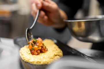 Chef plating tomato herb relish over creamy polenta on black plate in professional kitchen. Close-up of gourmet food preparation with stainless steel bowl in background.