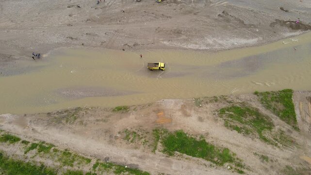 Aerial view of sand mining activity in Aileu, Timor-Leste. Trucks load aggregate from a riverbed extraction site next to green rice paddies, highlighting land use contrast.